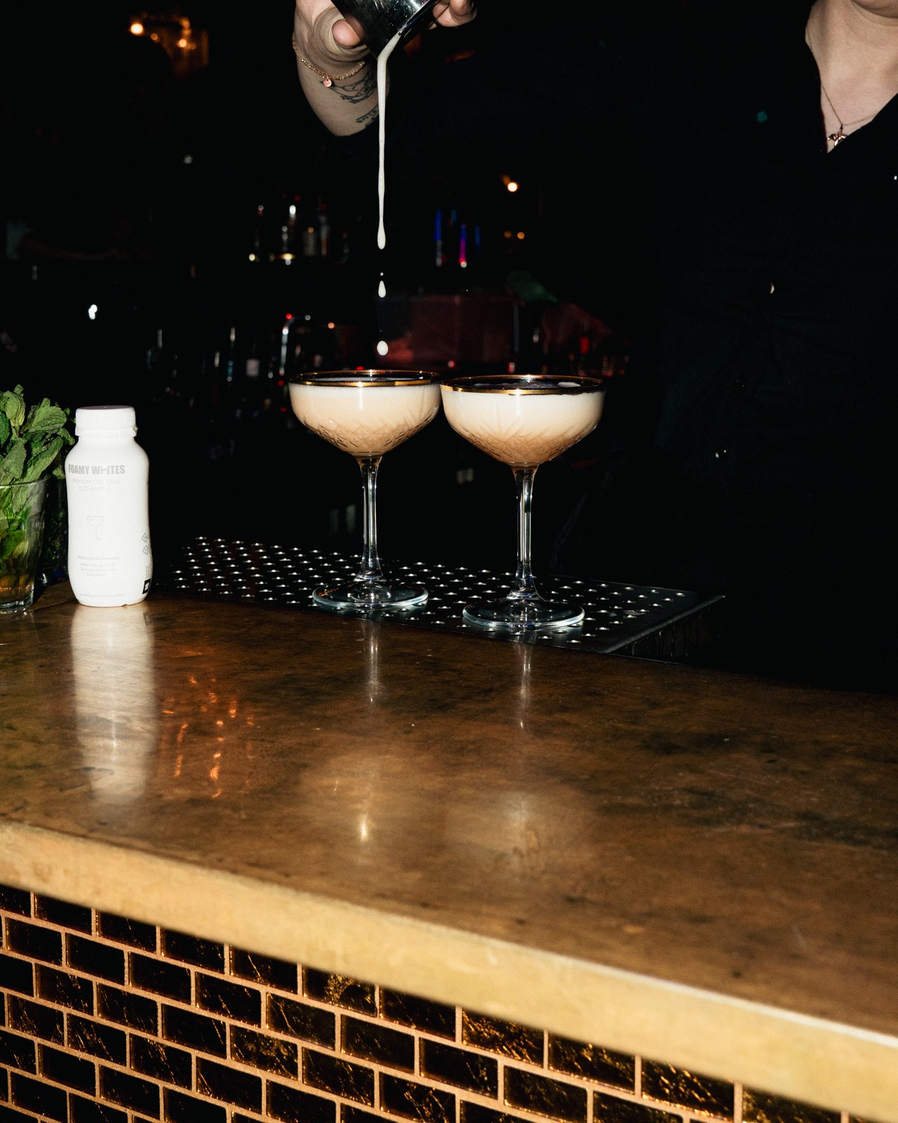 A bartender pours an egg-white cocktail beside a FOAMY WHITES bottle on a low-lit bar